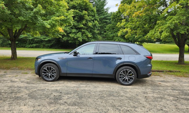 2025 Mazda CX-70 PHEV left profile view parked on gravel framed by trees.